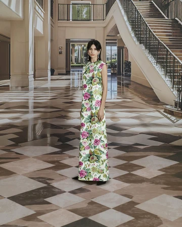 A woman in a floral dress stands in an indoor location with tile flooring, with stairs and windows in the background. 