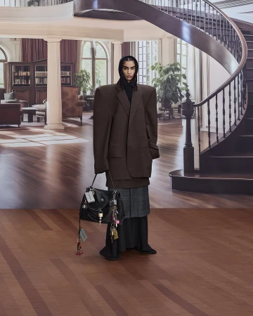 A female fashion model poses indoors wearing an oversized brown blazer, plaid skirt, black headscarf, and holding a black leather handbag.