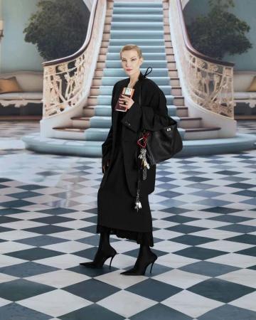 A woman in a black coat and skirt stands in front of a grand staircase with a handbag and book, photographed in a staged indoor setting.
