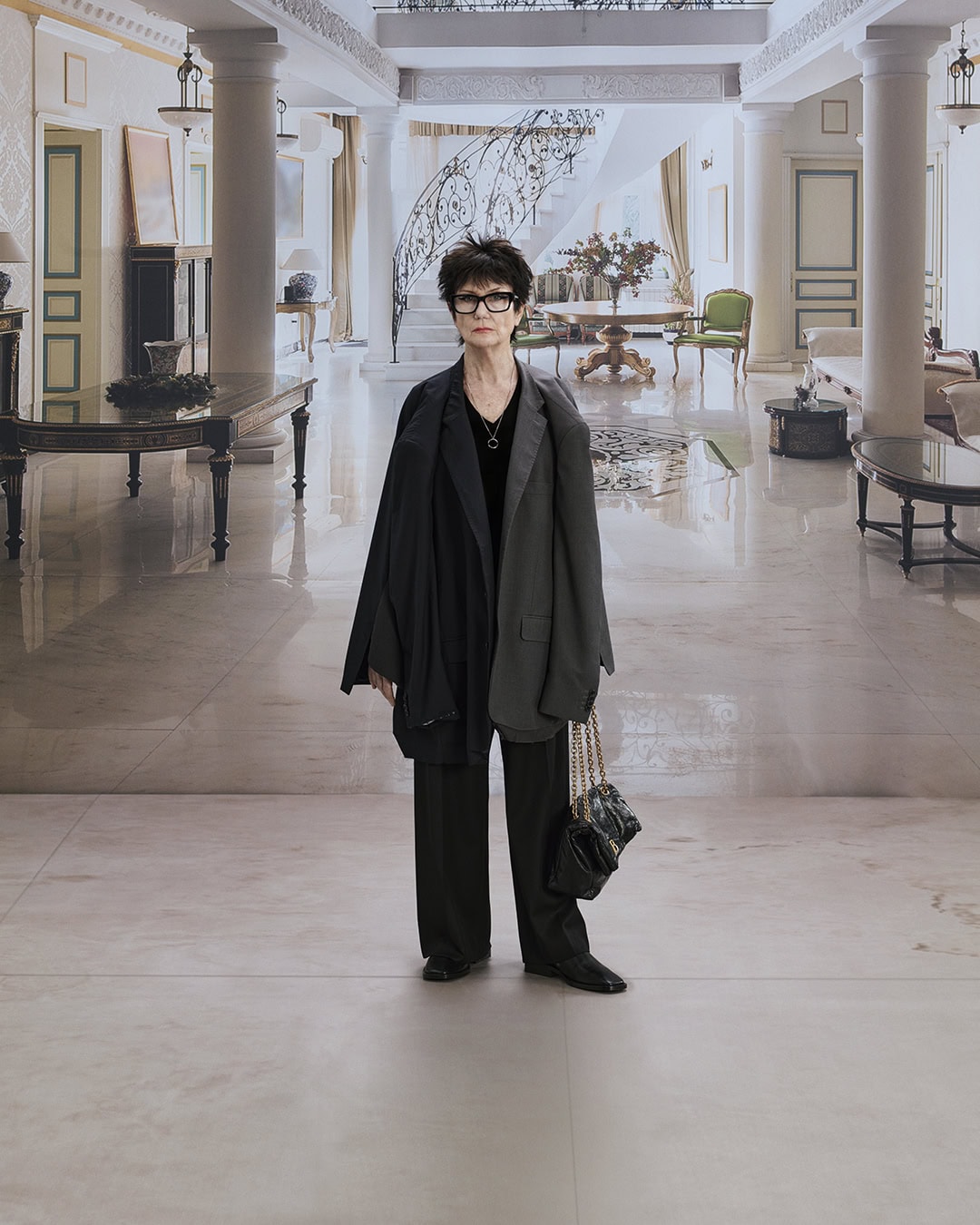 Middle-aged woman with glasses, wearing a blazer and carrying a purse, stands in a hallway with columns and marble floors.