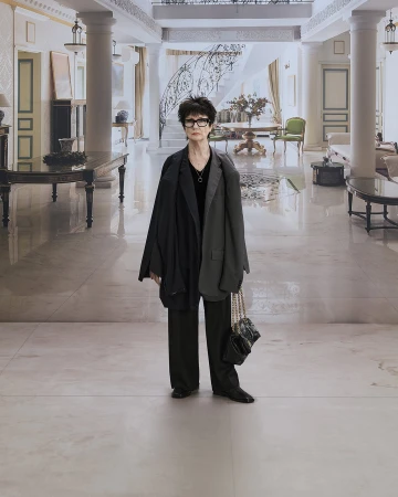 Middle-aged woman with glasses, wearing a blazer and carrying a purse, stands in a hallway with columns and marble floors.