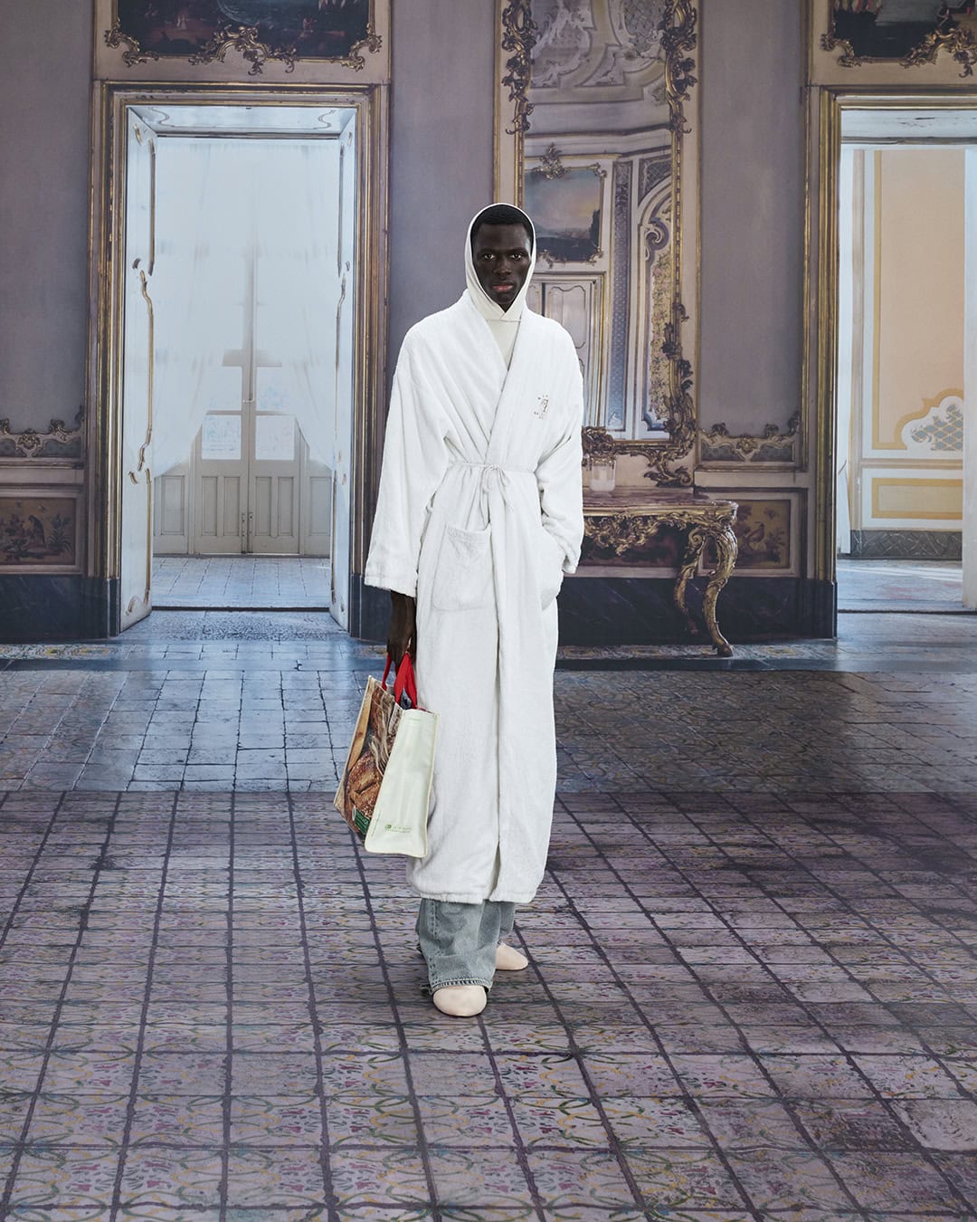 Fashion shot of an adult male wearing a white robe, carrying patterned shopping bags, posing indoors on a tiled floor in ornate hall.