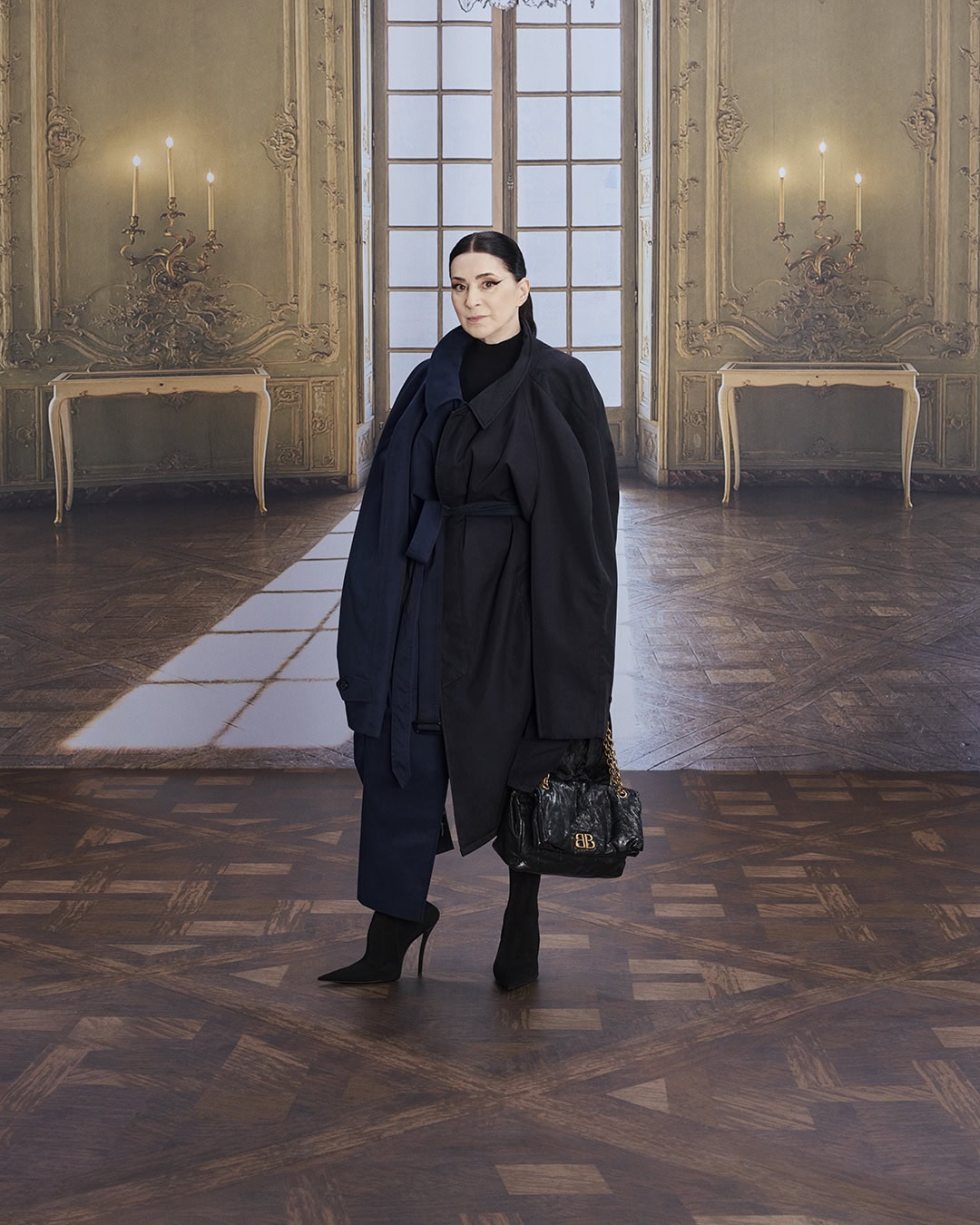 Elegant woman in a dark coat and heels stands in a grand hall with detailed walls, chandeliers, and a herringbone-patterned wood floor.