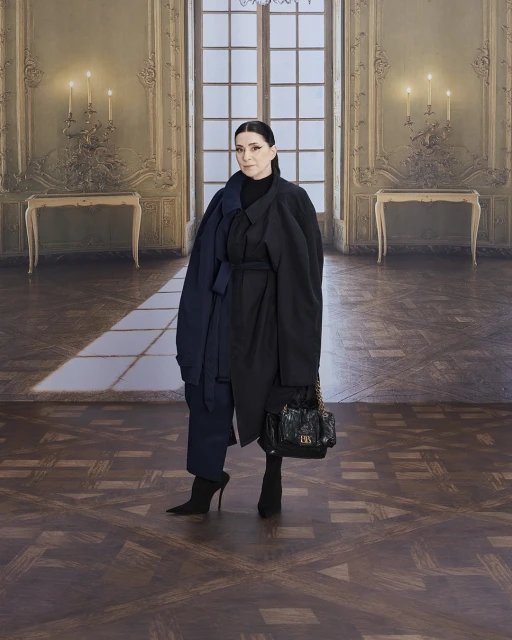 Elegant woman in a dark coat and heels stands in a grand hall with detailed walls, chandeliers, and a herringbone-patterned wood floor.