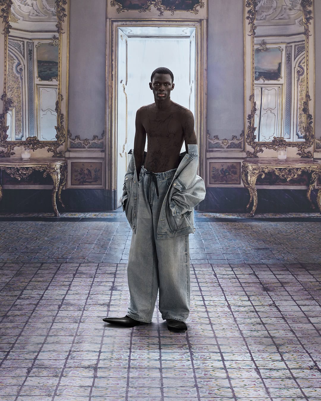 A shirtless African-American adult male stands in an ornately decorated room wearing oversized denim pants and a jacket over his shoulders.