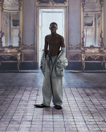 A shirtless African-American adult male stands in an ornately decorated room wearing oversized denim pants and a jacket over his shoulders.