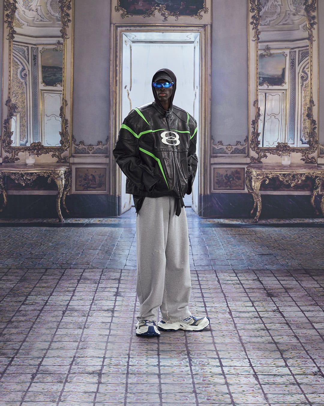 An adult fashion model poses in a black and green jacket and gray sweatpants in a large indoor room with an ornate architectural design.
