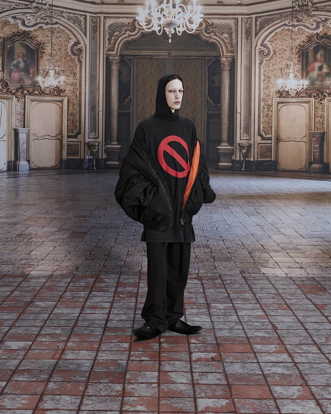 Full shot of an adult wearing a black hooded shirt with a red no symbol, black jacket, and black pants, in an ornate room.