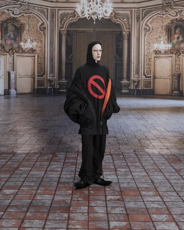Full shot of an adult wearing a black hooded shirt with a red no symbol, black jacket, and black pants, in an ornate room.