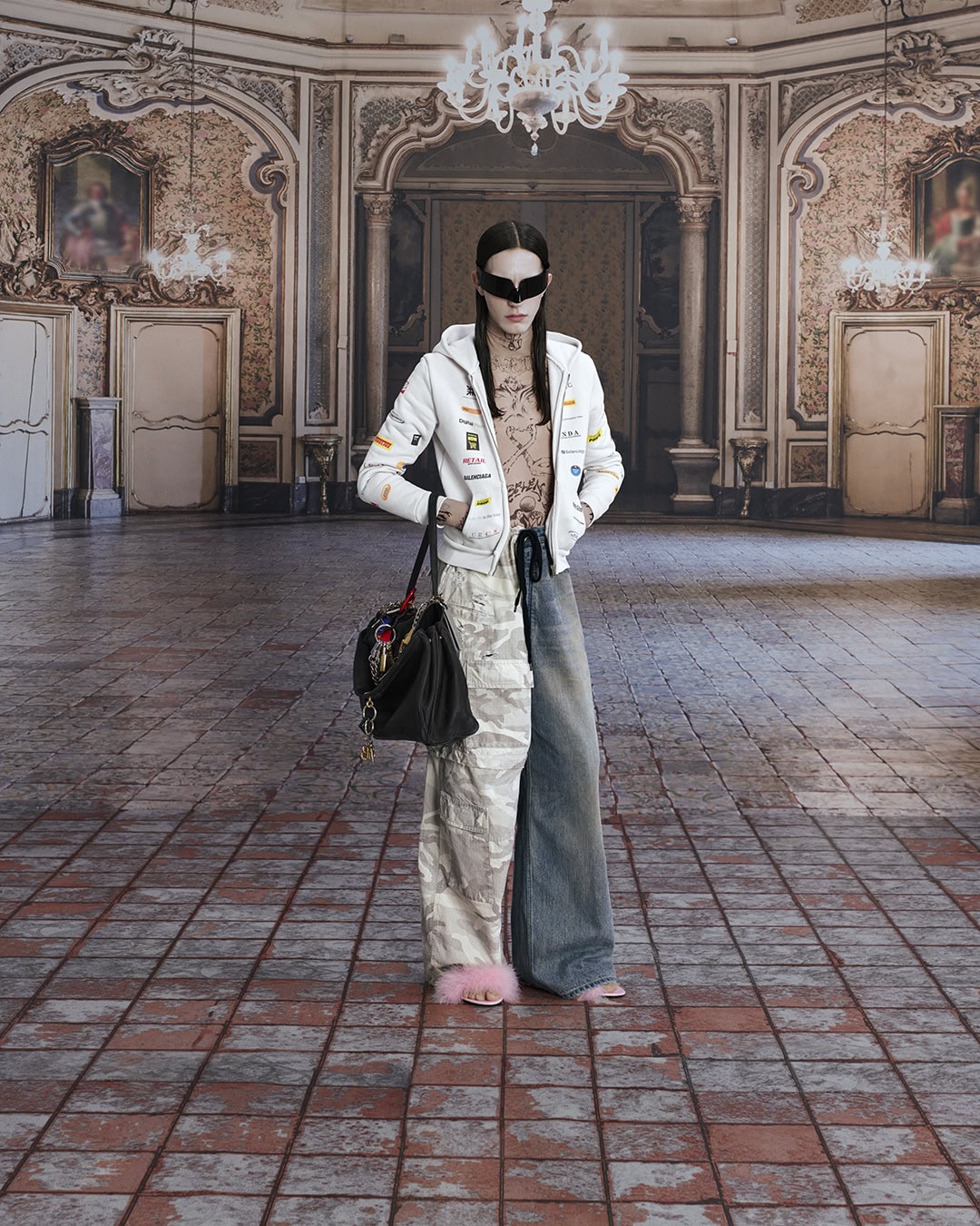 A person stands in a grand, ornate room wearing an eclectic outfit including a white hoodie, mismatched jeans, and pink fuzzy slippers.