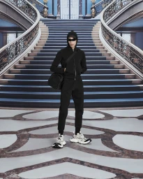 A masked model in a black hoodie and sweatpants holding a black crossbody bag stands before a grand staircase.