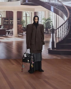 Model wearing an oversized brown blazer and carrying a large black handbag standing in an interior salon setting