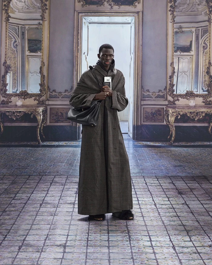 Model wearing a long patterned coat and carrying a black leather handbag inside an ornate room with large mirrors and tiled floor.