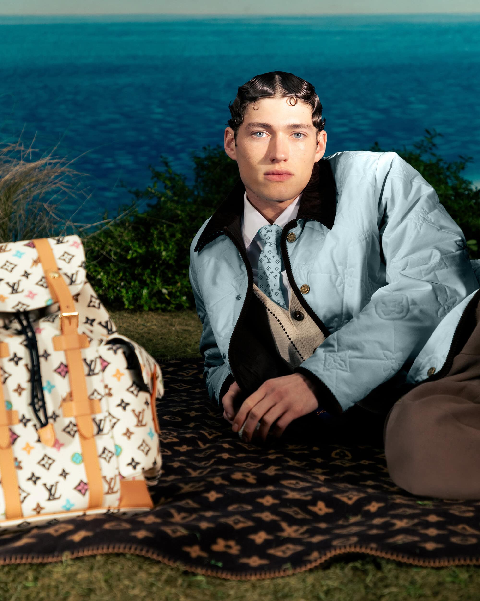 A fashionable man poses outdoors on a patterned blanket next to a Louis Vuitton backpack, with a blue sea in the background.
