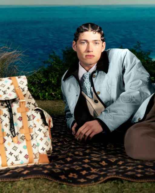 A fashionable man poses outdoors on a patterned blanket next to a Louis Vuitton backpack, with a blue sea in the background.
