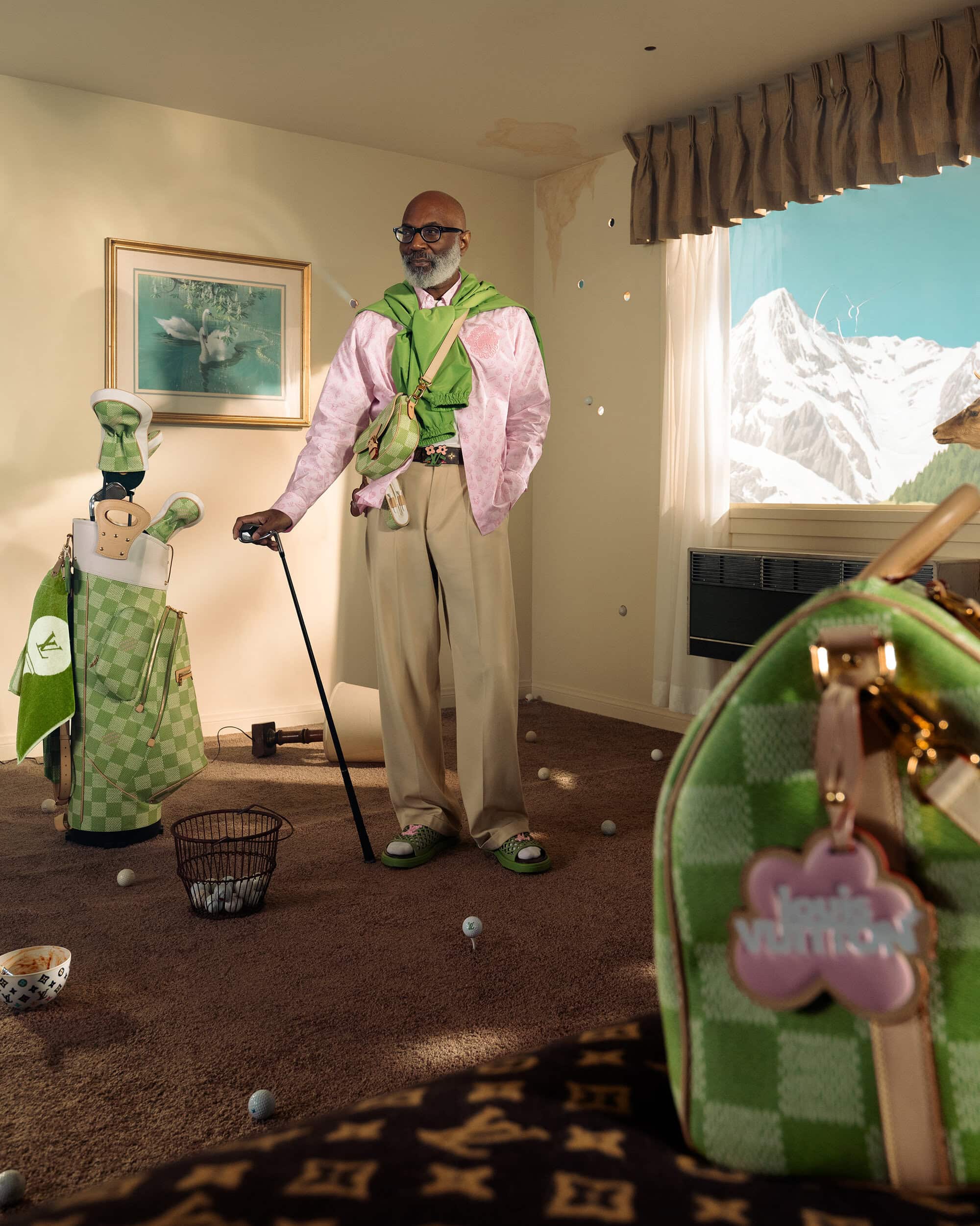 Man in pink jacket holding golf club in quirky indoor golf scene with golf balls, bag, and mountain view through window.