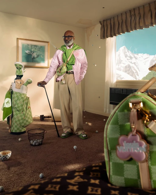 Man in pink jacket holding golf club in quirky indoor golf scene with golf balls, bag, and mountain view through window.