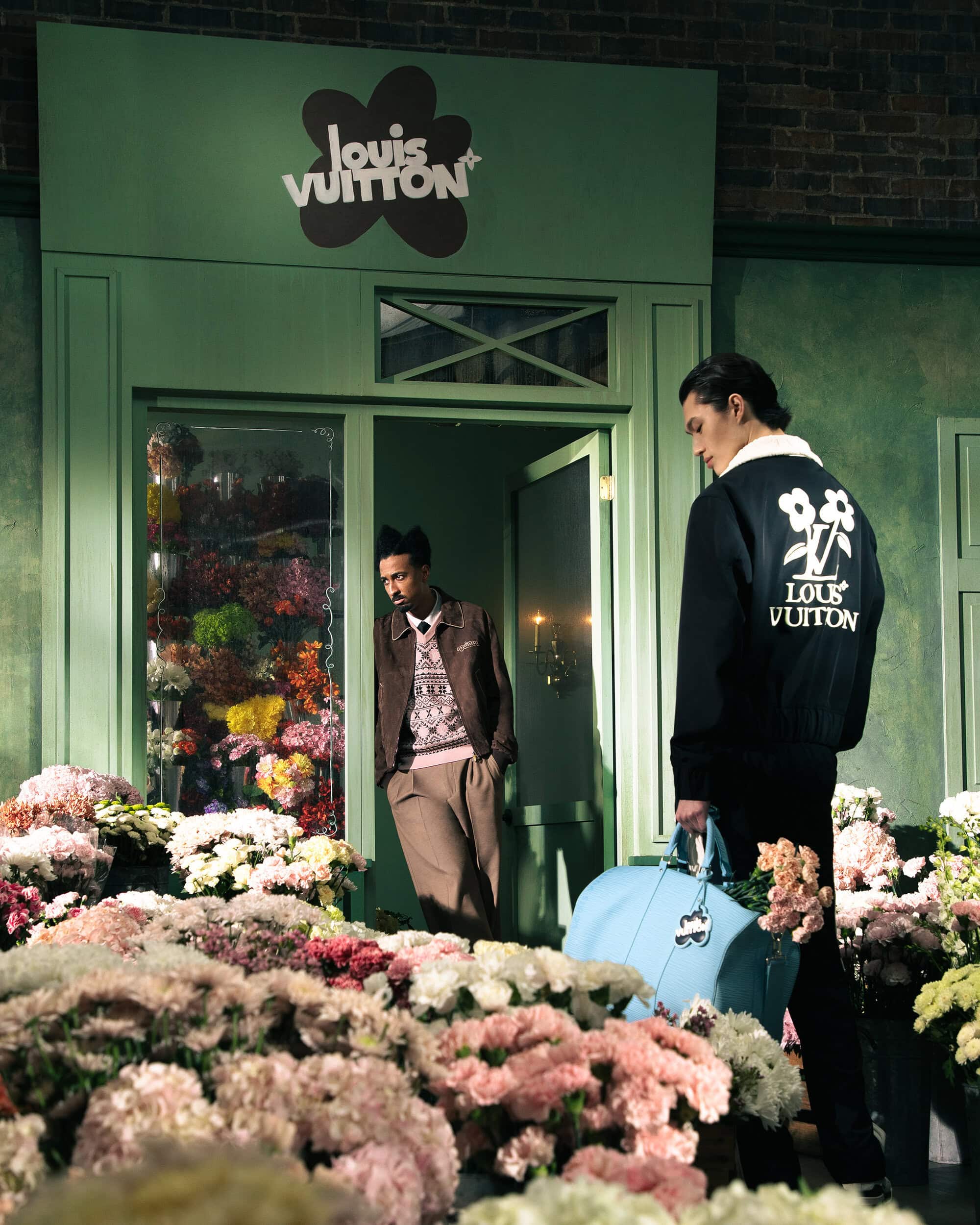 Two men stand near a flower-filled shop entrance with a Louis Vuitton sign; one man wears a branded jacket, carrying a light-blue duffel bag.
