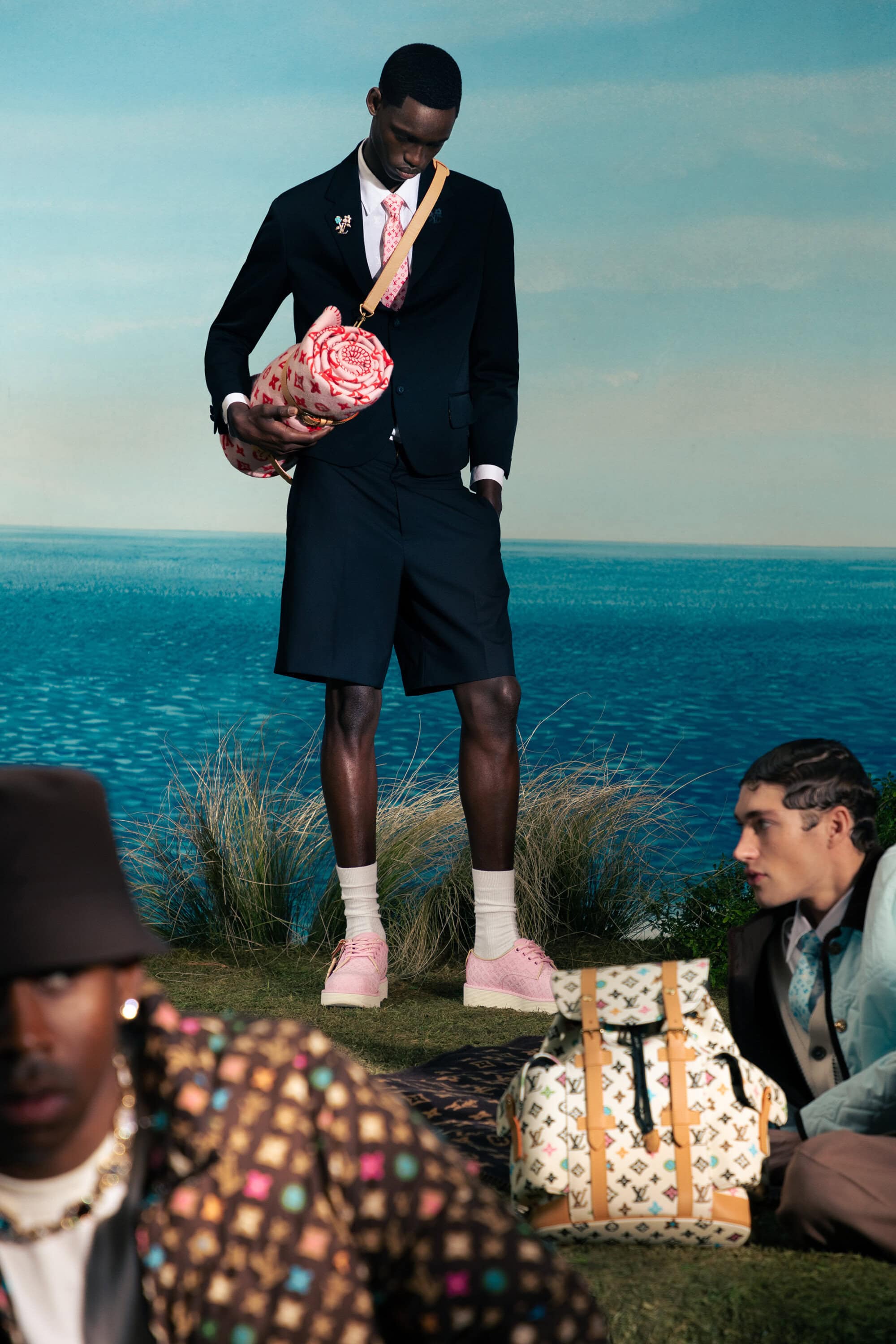 Three men model the Louis Vuitton 2023 pre-collection, wearing jackets, shorts, pastel shoes and carrying patterned bags with an ocean backdrop.