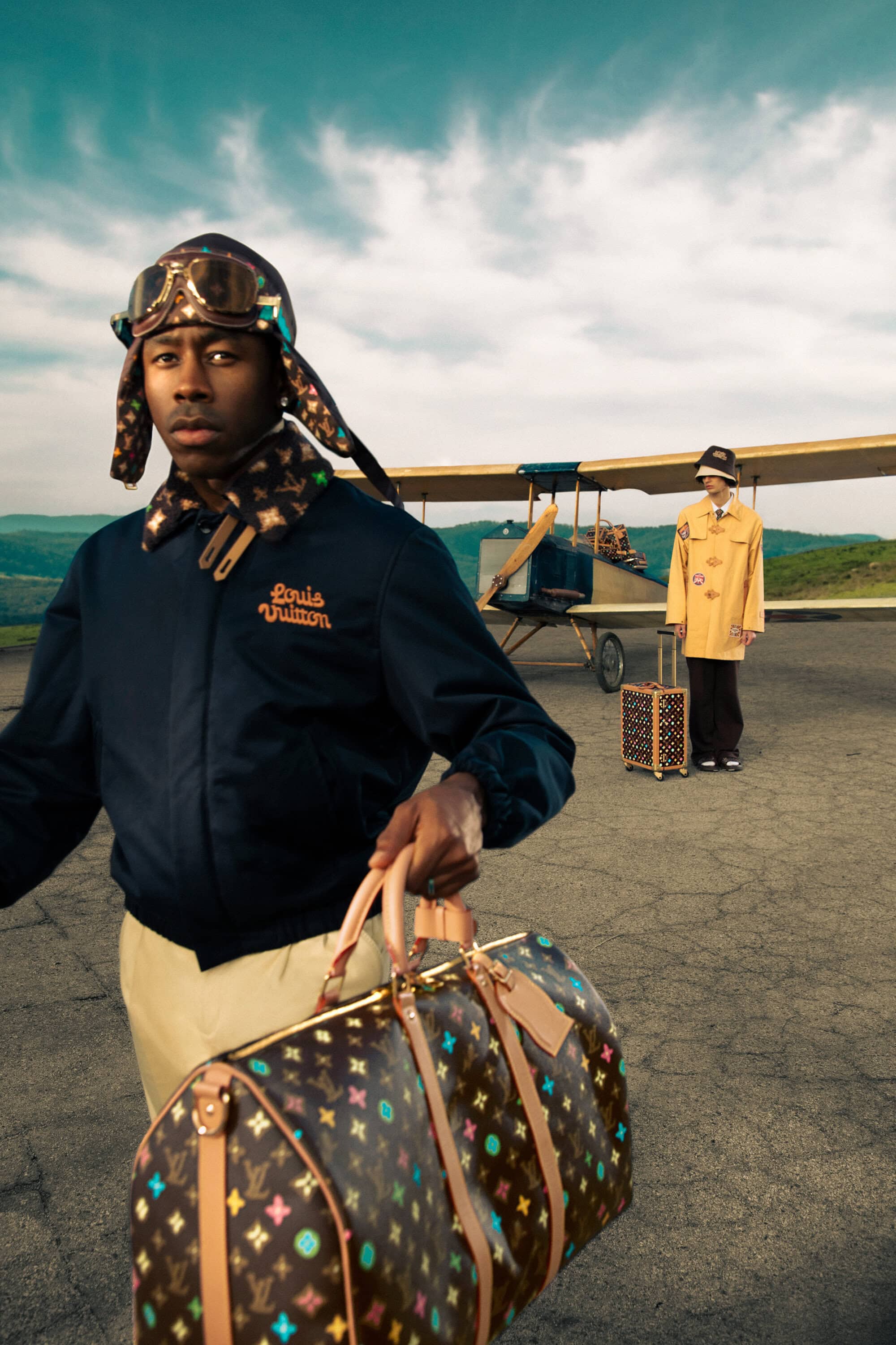 Tyler the Creator stands with a Louis Vuitton bag and aviator hat near a vintage airplane on an airstrip, fashion editorial.