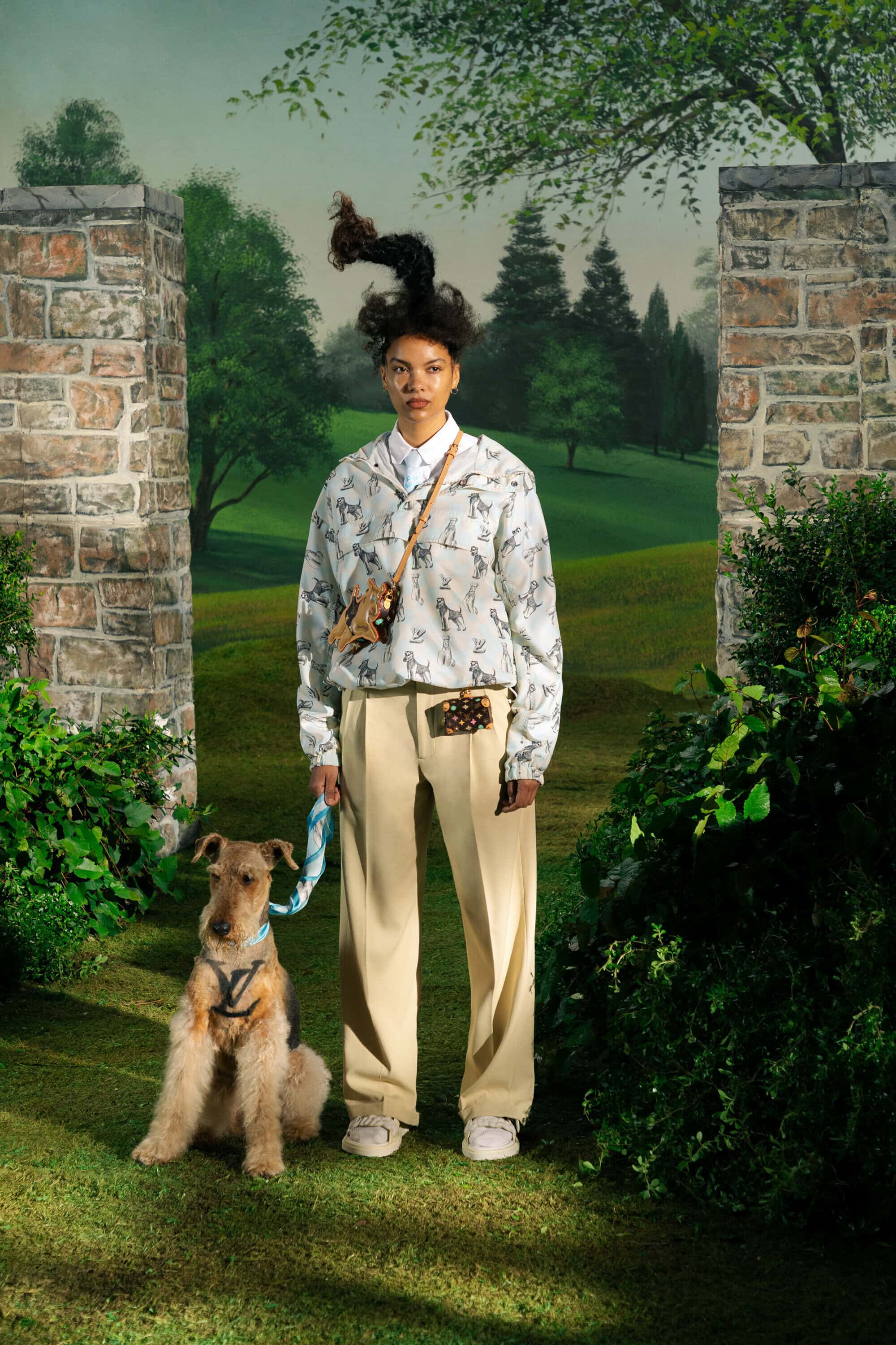 A young adult woman poses with an Airedale Terrier outdoors on a green lawn in front of a painted landscape backdrop.