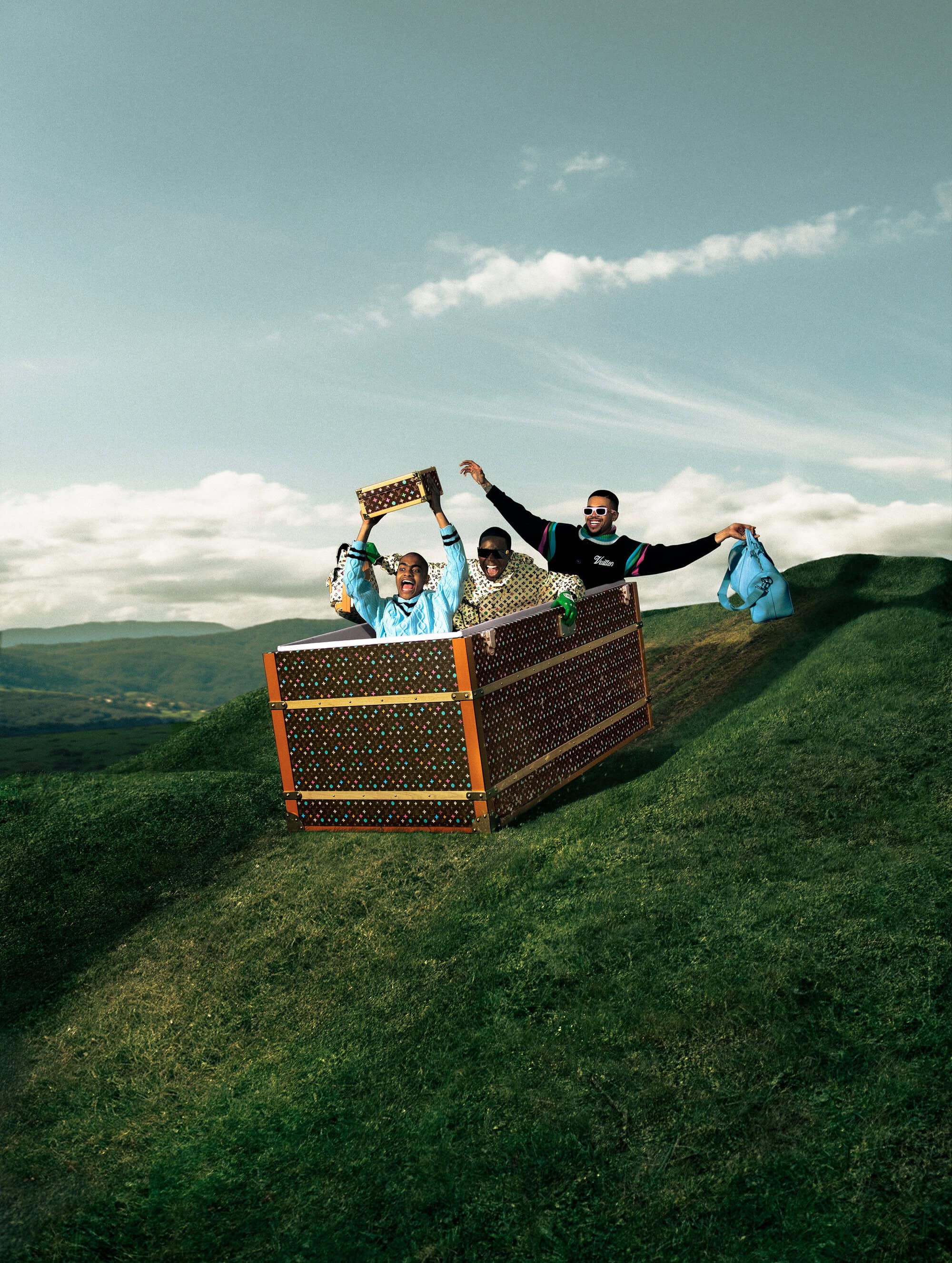 Three adults joyfully ride in a Louis Vuitton trunk downhill on a grassy hill under a partly cloudy blue sky in an outdoor setting.