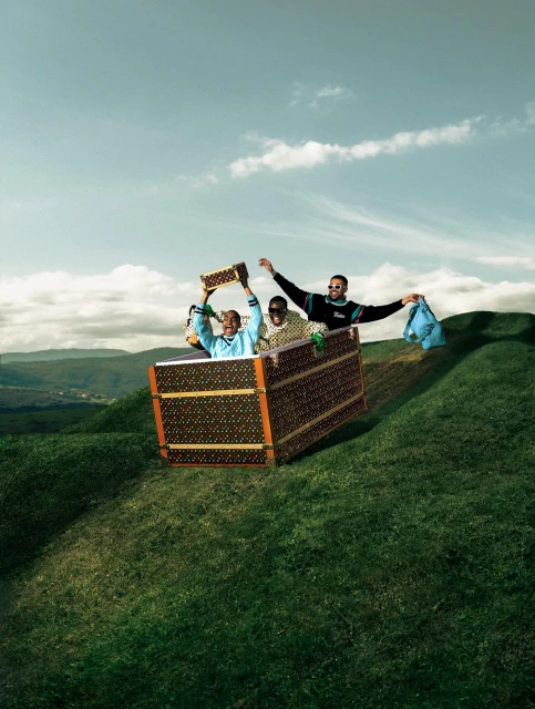 Three adults joyfully ride in a Louis Vuitton trunk downhill on a grassy hill under a partly cloudy blue sky in an outdoor setting.