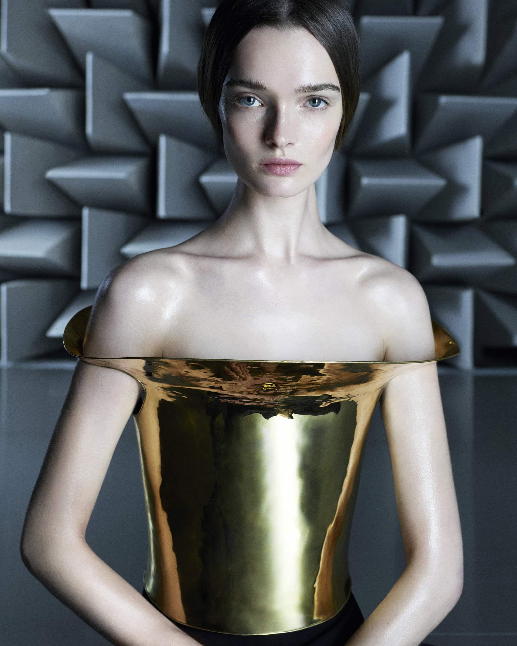 Young woman poses in a golden metal corset against a geometric background in this fashion-forward studio portrait.