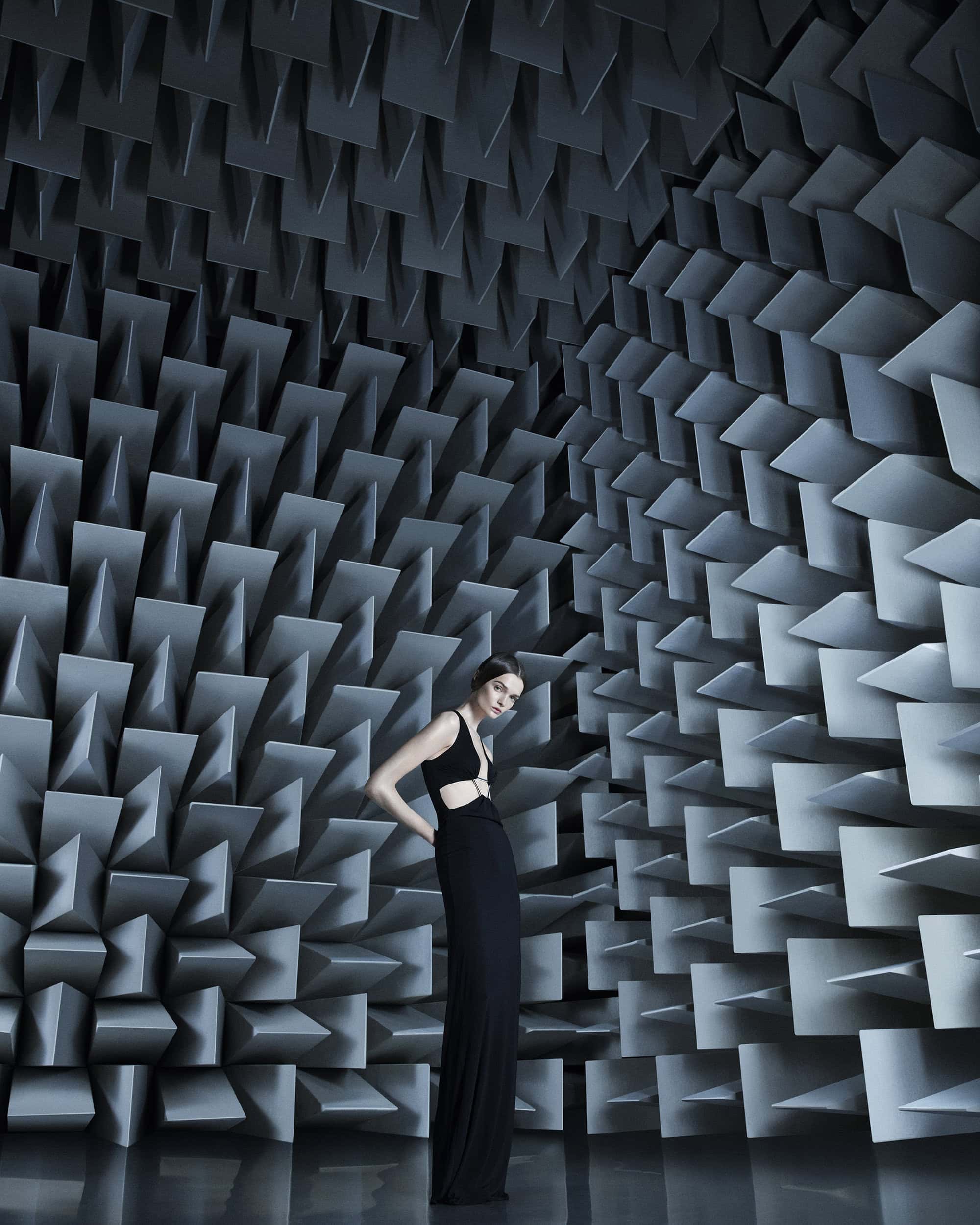 Woman in a black dress poses in an anechoic chamber with gray soundproofing wedges covering the walls and ceiling, reflected in the shiny floor.