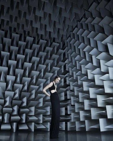 Woman in a black dress poses in an anechoic chamber with gray soundproofing wedges covering the walls and ceiling, reflected in the shiny floor.