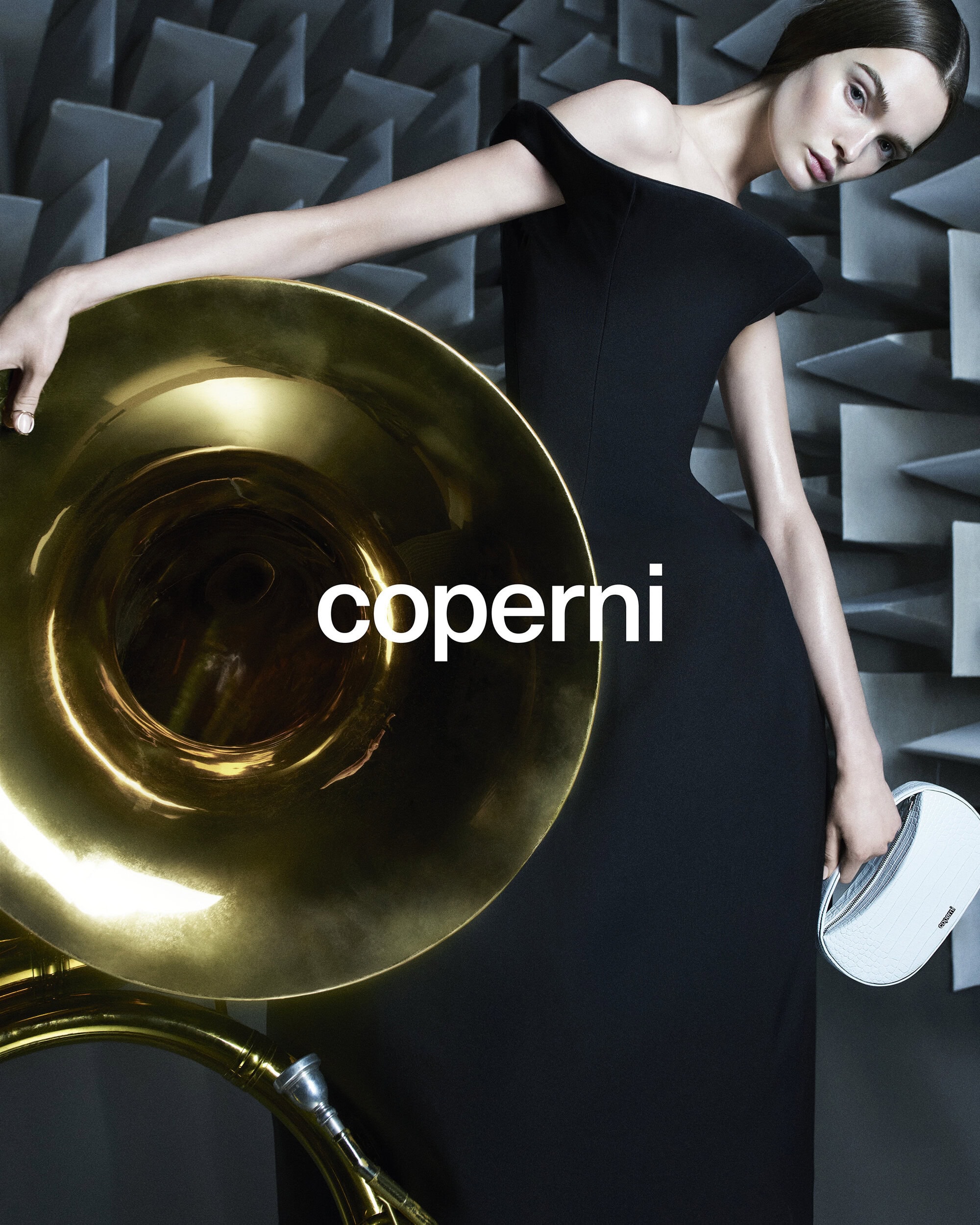 A woman in an elegant black dress poses with a tuba and a white hard case against an abstract grey geometric background indoors.