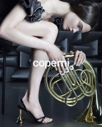 Model seated wearing black dress and heels holding a French horn for Coperni Spring 2024 campaign