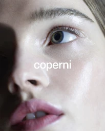 Extreme close-up portrait of a woman's face with light makeup and a prominent white Coperni logo across the bridge of her nose.