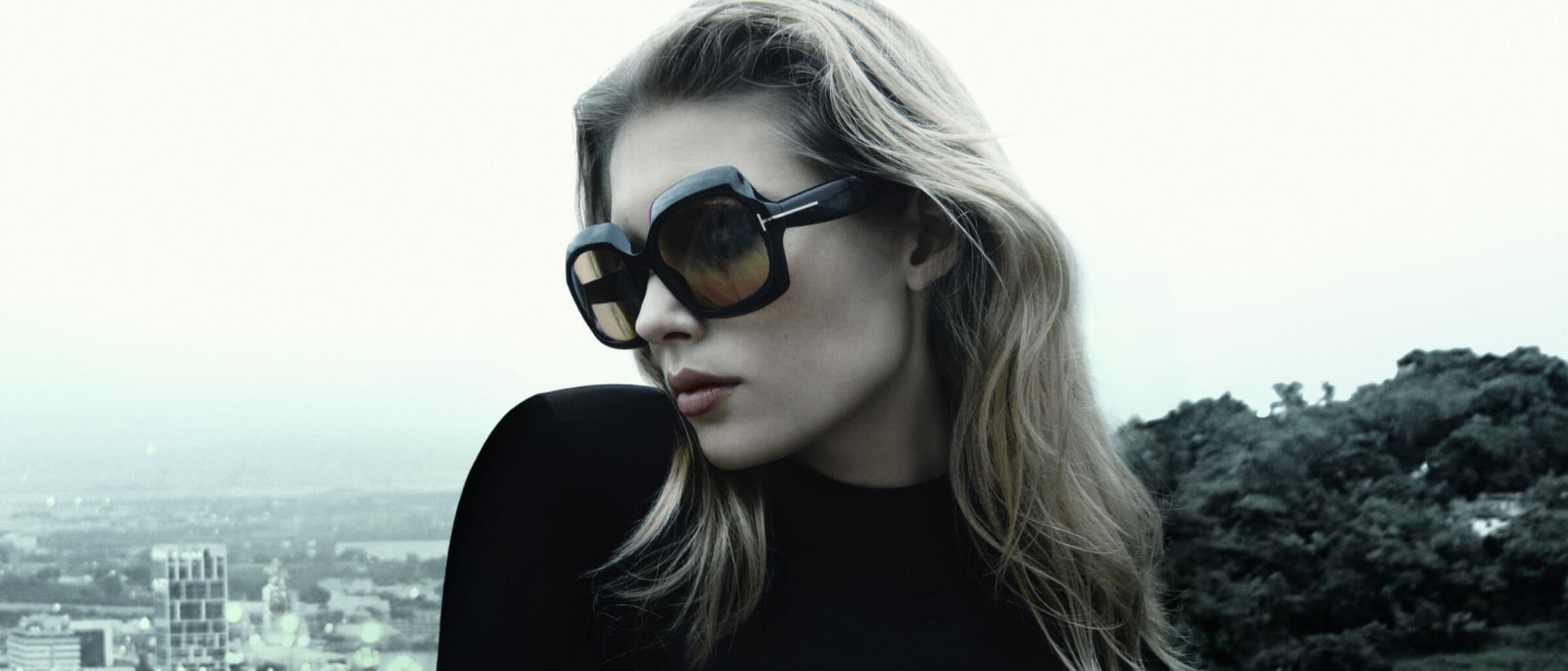 Close-up of a blonde woman wearing oversized black sunglasses and a black top, posing in front of a cityscape and vegetation.