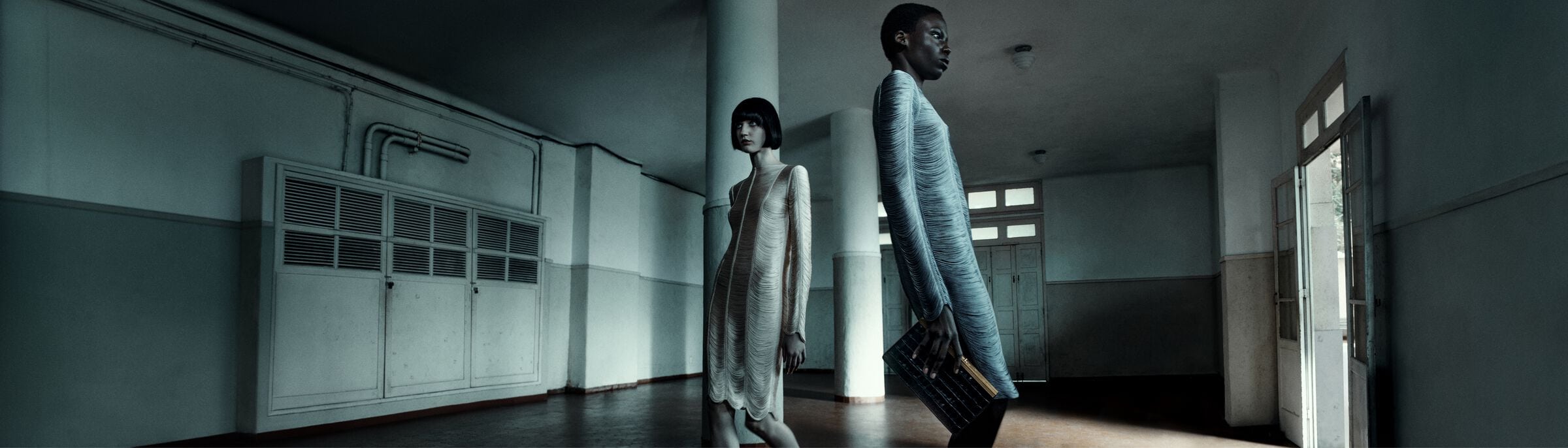 Two women wearing light-colored dresses stand in a long, empty corridor with white walls, pillars, and doors in a cool-toned fashion shot.