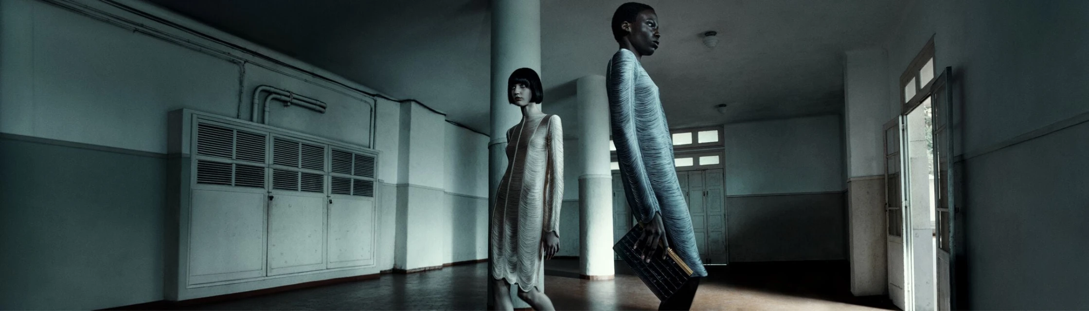 Two women wearing light-colored dresses stand in a long, empty corridor with white walls, pillars, and doors in a cool-toned fashion shot.