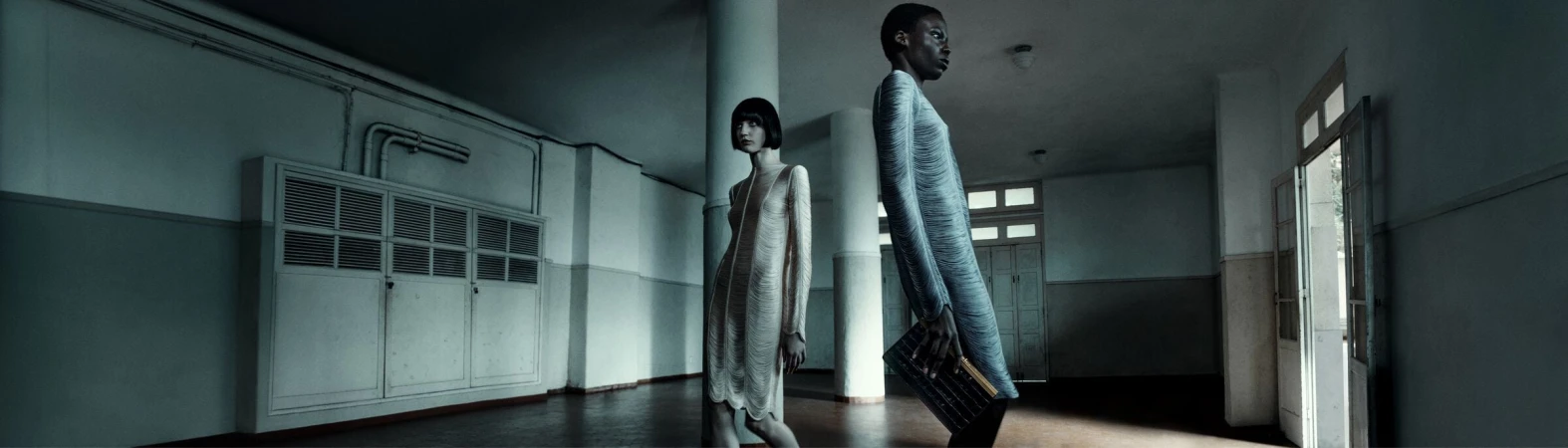 Two women wearing light-colored dresses stand in a long, empty corridor with white walls, pillars, and doors in a cool-toned fashion shot.