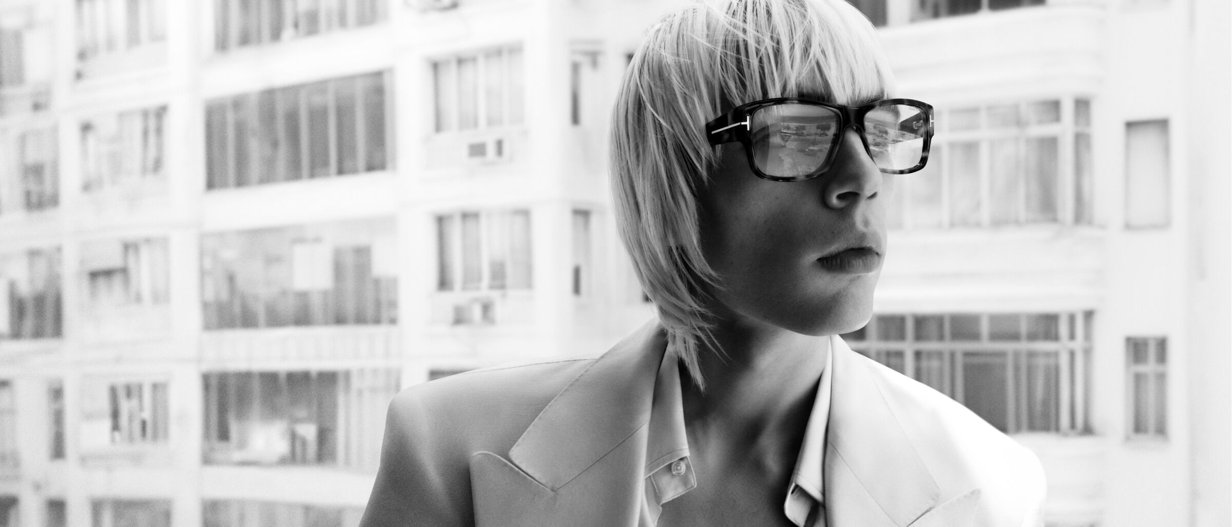 Black and white portrait of a blonde person with glasses, wearing a white jacket, against a blurred building backdrop.