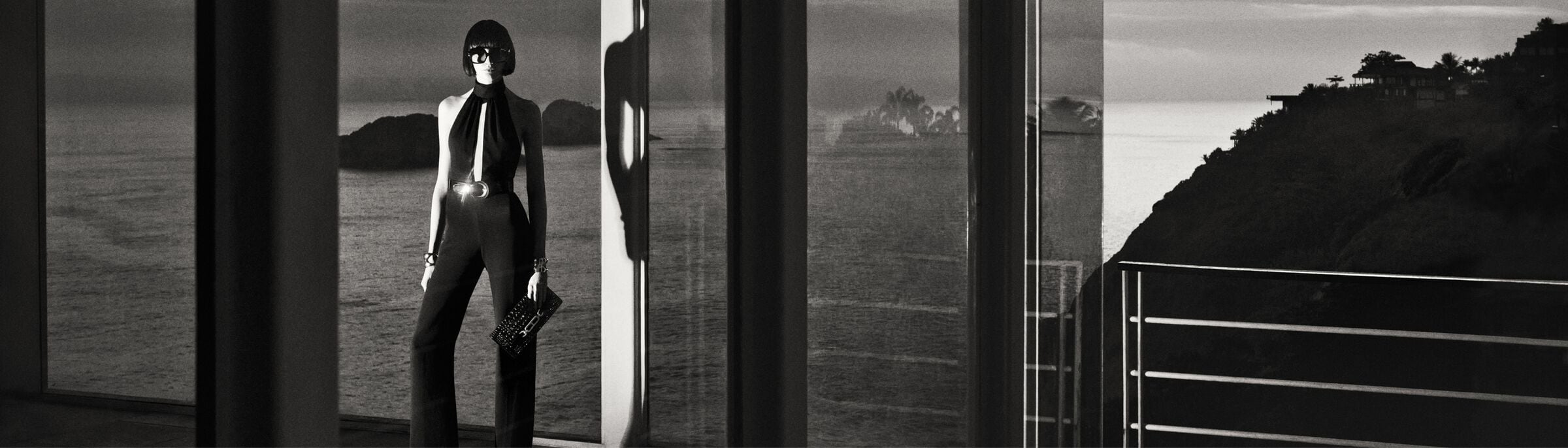 Black and white fashion shot of an adult female model in a halter jumpsuit holding a clutch, posing in front of a large window.