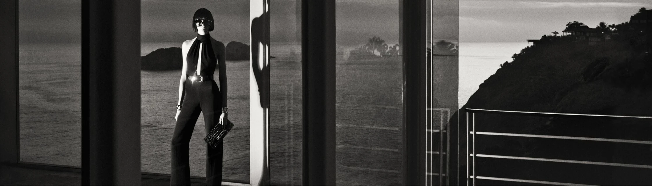 Black and white fashion shot of an adult female model in a halter jumpsuit holding a clutch, posing in front of a large window.