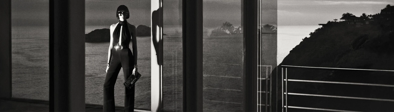 Black and white fashion shot of an adult female model in a halter jumpsuit holding a clutch, posing in front of a large window.