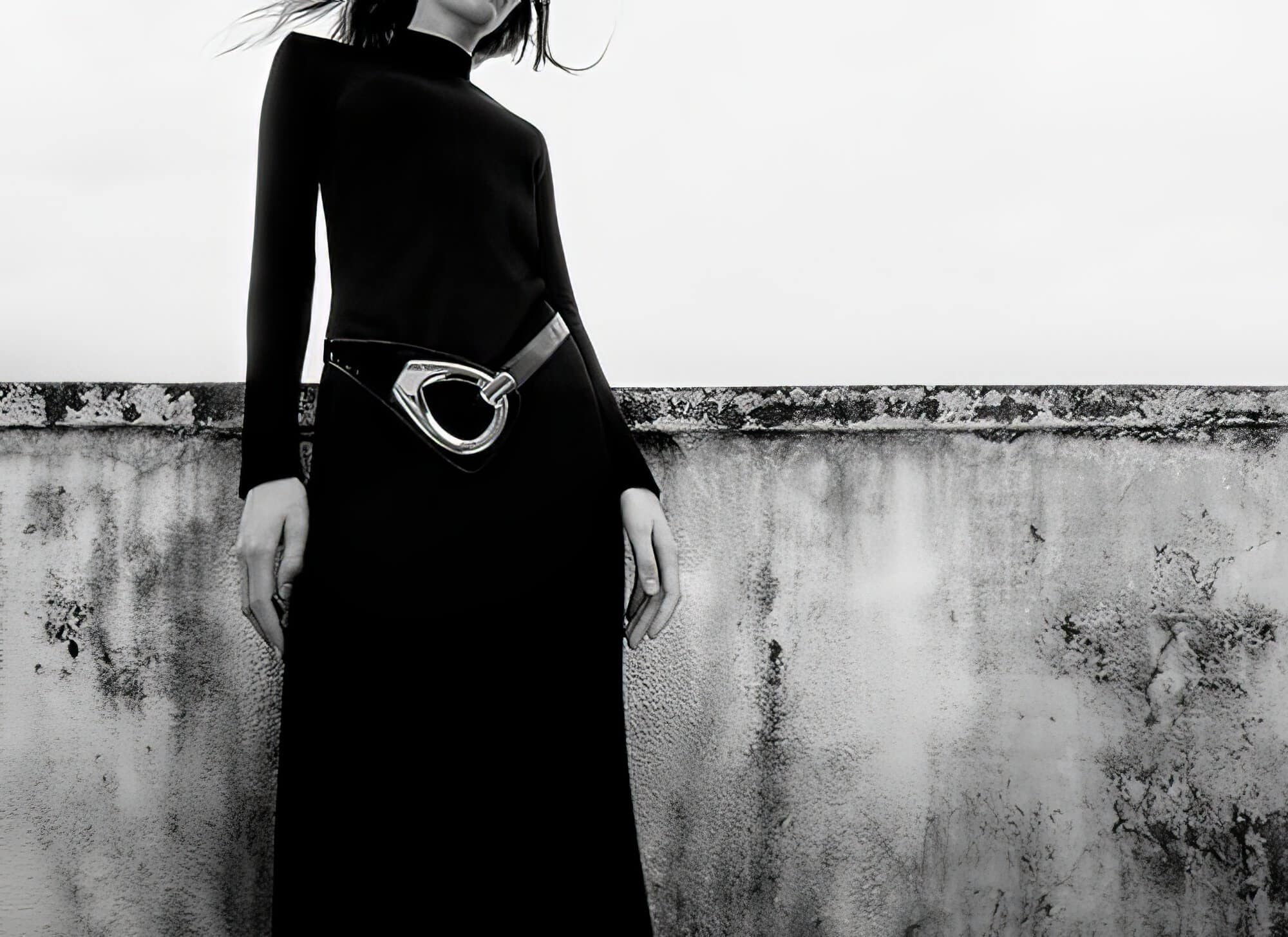 A woman in a long black dress poses against a textured wall in a black and white fashion shot with muted, neutral tones.