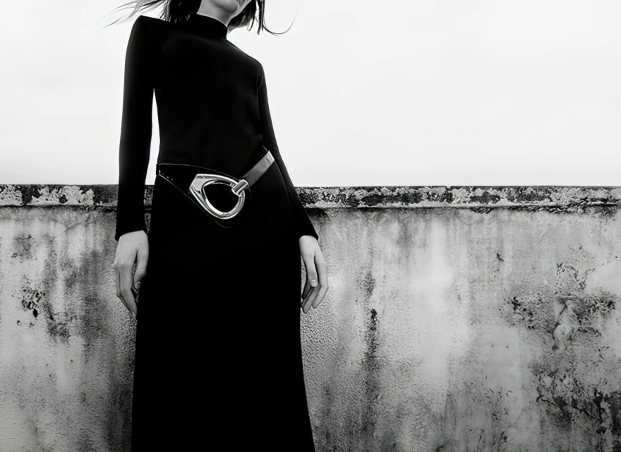 A woman in a long black dress poses against a textured wall in a black and white fashion shot with muted, neutral tones.
