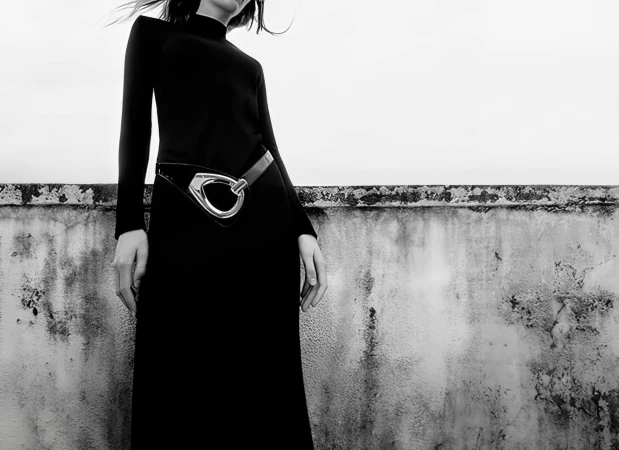 A woman in a long black dress poses against a textured wall in a black and white fashion shot with muted, neutral tones.