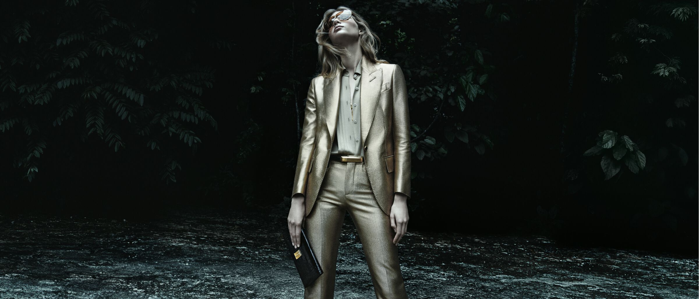 Fashionable woman posing in a shiny gold suit outdoors. She is wearing sunglasses and holding a black purse. Dark foliage background.