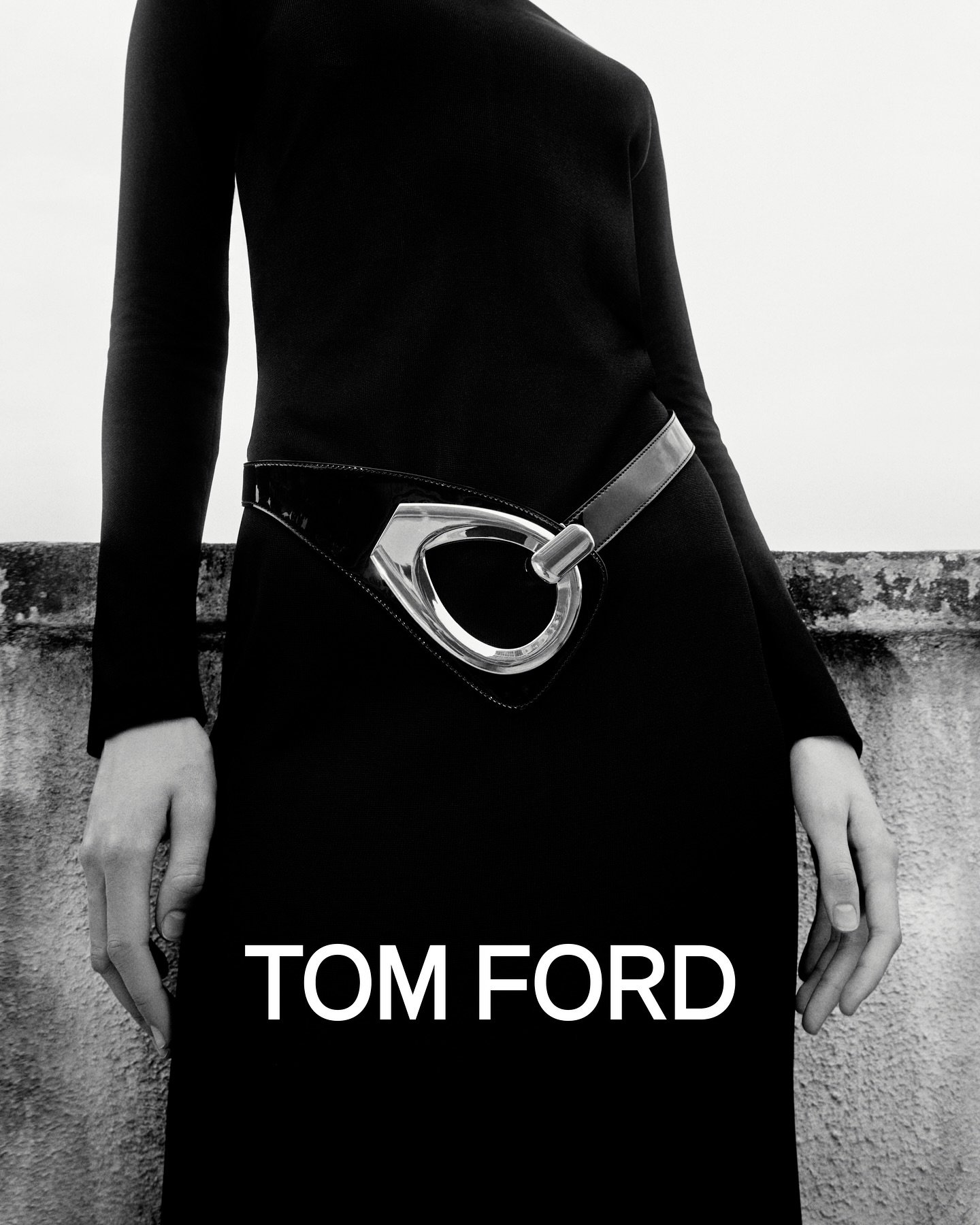 A fashion shot of a model wearing a black dress with a striking silver belt, set against a textured wall and a bright white sky.