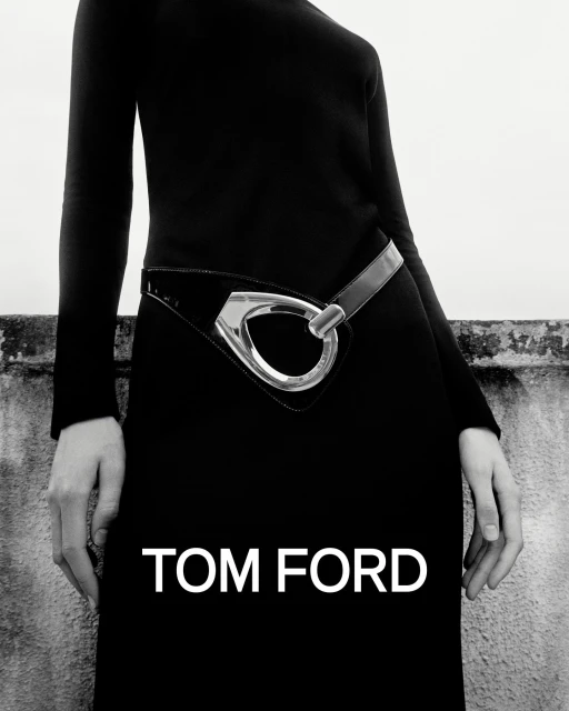 A fashion shot of a model wearing a black dress with a striking silver belt, set against a textured wall and a bright white sky.