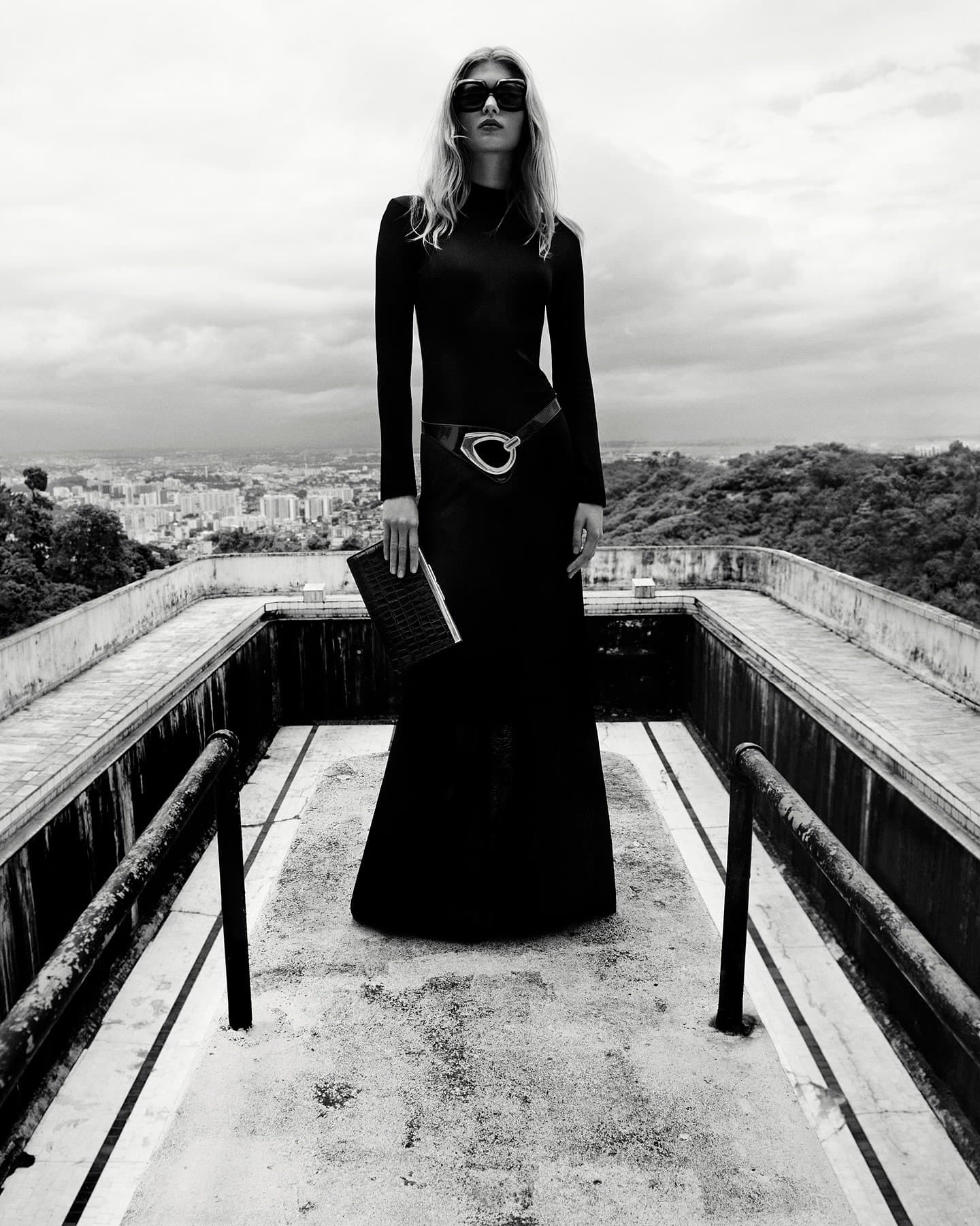 Monochrome fashion portrait of a woman in a long black dress and sunglasses, posing on a rooftop overlooking a cityscape.