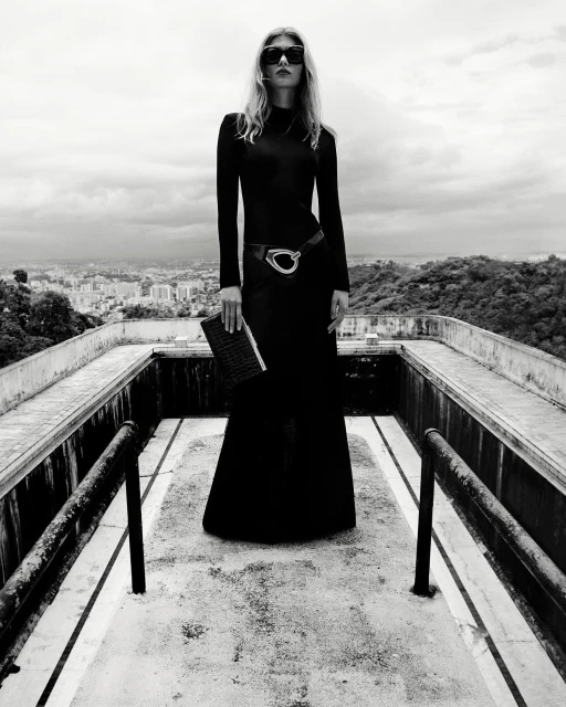 Monochrome fashion portrait of a woman in a long black dress and sunglasses, posing on a rooftop overlooking a cityscape.