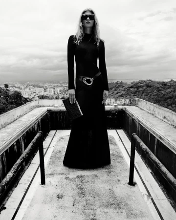 Monochrome fashion portrait of a woman in a long black dress and sunglasses, posing on a rooftop overlooking a cityscape.